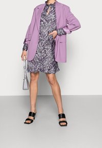 Fioletowa marynarka nałożona na wzorzystą fioletową sukienkę z falbanistym kołnierzykiem, w połączeniu z fakturowanymi czarnymi szpilkami i małą szaro-skalną torebką.
