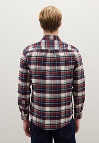 Camisa de hombre a cuadros en azul marino, rojo y crema. Mangas largas, cuello, puños con botones y dobladillo curvo; confeccionada en algodón con una textura suave.