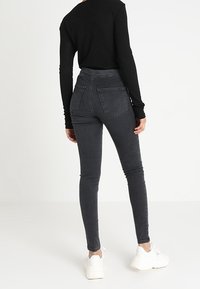 Osoba oblečená v černém dlouhém rukávu, tmavě šedých skinny džínách a bílých robustních teniskách, stojící zády k bílé stěně.