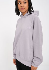 Haut à capuche violet clair avec une coupe ample, capuche à cordon, poignets et ourlet côtelés. Comprend un petit logo brodé sur la poitrine.