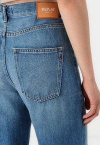 Lichtblauwe denimjeans met een brown leren label met de tekst "REPLAY" op de tailleband. Heeft zichtbare stiksels en een achterzak met een embleem.