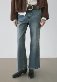 Wijde jeans in medium blauwe denim met een gladde textuur, gecombineerd met een bruine suède jas en zwarte enkellaarsjes met hak.