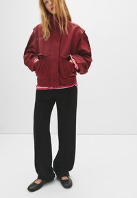 Burgundska oversized jakna s velikim džepovima i stojećim kragnom, složena preko crveno-bijele prugaste košulje, u kombinaciji s crnim širokim hlačama.