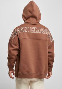 Pruun kapuutsiga sviiter, mille seljal on suur valge "URBAN CLASSICS" tekst. Valmistatud pehmest materjalist, omab kapuutsi ja mugavat lõiget. Nähtavad õmblused.