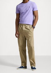 Man som står klädd i en lavendelfärgad t-shirt med kort ärm, beige veckade byxor, blå kanvas sneakers och ett armbandsur, med händerna i fickorna.