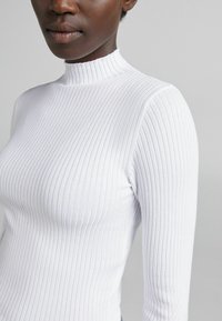 Haut col roulé blanc côtelé à manches longues, avec un col montant et une texture subtile, silhouette ajustée avec une finition lisse.