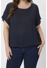 Femme portant un blouse à manches courtes bleu marine et un jean bleu foncé, se tenant avec une main dans la poche devant un fond clair uni.