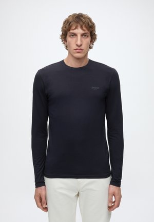 Giovane uomo con capelli ricci che indossa una maglia nera aderente a maniche lunghe e pantaloni bianchi, in piedi davanti a uno sfondo chiaro e uniforme.