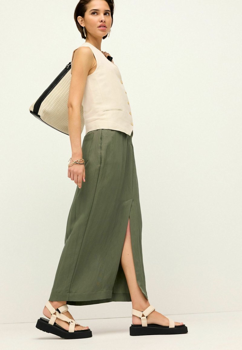 Top en crème à bretelles avec des boutons, jupe maxi olive verte avec une fente sur le côté, et sandales à lanières beiges sur un modèle, tenant un sac texturé beige.