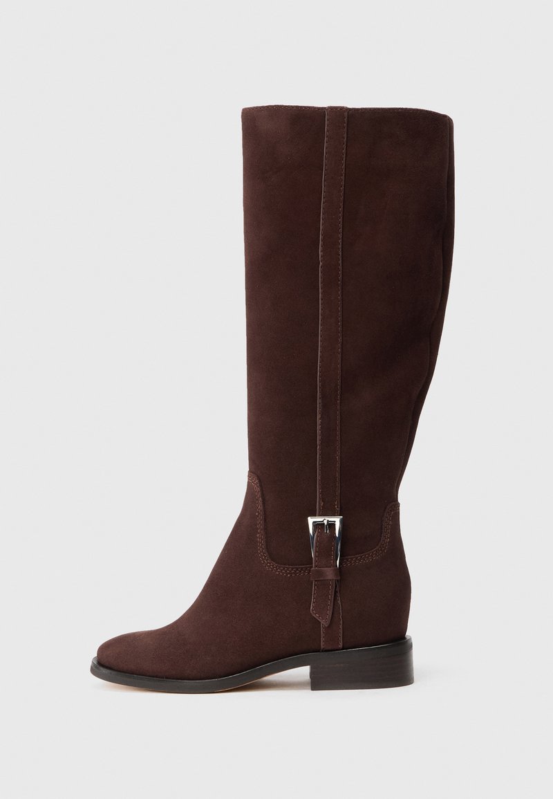 Bota de camurça castanha até ao joelho, com biqueira quadrada, salto plano e um detalhe decorativo de fivela lateral. Textura suave com acabamentos cosidos.