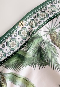 Tkanina o tropikalnym wzorze przedstawiająca liście palmowe i ananasy w odcieniach zieleni. Akcentowana ciemnozielonym obszyciem i złotymi elementami ozdobnymi.