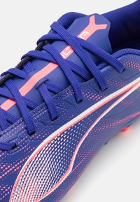 Zapato de fútbol sintético azul con acentos rosas, superficie texturizada y detalles en blanco contrastantes. Presenta cordones prominentes y un diseño curveado.