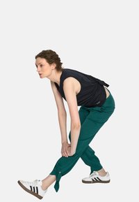 Młoda kobieta w czarnej bezrękawnikowej bluzce i zielonych spodniach rozciąga lewą nogę, trzymając kostkę rękami, w białych sneakersach Adidas.