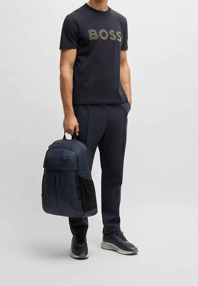 Mies, joka on pukeutunut mustaan "BOSS"-teepaitaan, mustiin housuihin ja lenkkikenkiin, pitää kiinni tummansinisestä reppusta, jonka etutaskussa on "BOSS"-logo.