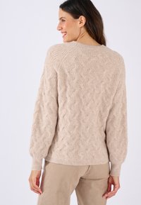Pull en tricot beige avec un motif de câble texturé, des poignets côtelés et un ourlet légèrement court, porté sur un pantalon en velours côtelé beige.