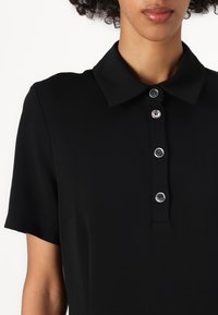 Polo shirt nera con colletto e maniche corte, caratterizzata da una fila di cinque bottoni argentati, texture liscia e design minimalista.