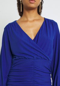 Blå omlottklänning i texturerad tyg, med långa  ärmar, djup v-ringning och samlad detalj vid midjan.