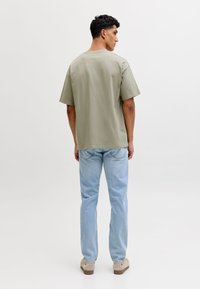 Vīrietis tērpts gaiši zaļā pārmēru plašā īsroku krekla, gaiši zilās džinsās un bēšās kurpēs, stāv ar muguru pret vienkrāsainu fonu.