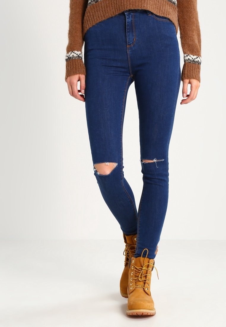 Skinny denim jeans in dark blue with distressed knee rips, paired with tan lace-up boots. The outfit features a ribbed brown sweater.