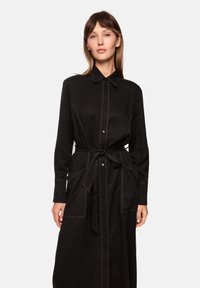 Schwarzes Hemdblusenkleid mit langen Ärmeln, einem Bindegürtel und kontrastierenden weißen Nähten. Verfügt über zwei vordere Taschen und einen klassischen Kragen.