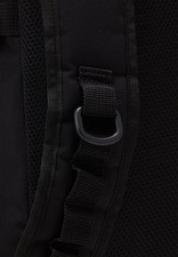 Borsa nera in nylon con pannello in rete, caratterizzata da una superficie texture, tracolla regolabile e un anello metallico circolare per il fissaggio. Dettaglio di cucitura regolare.
