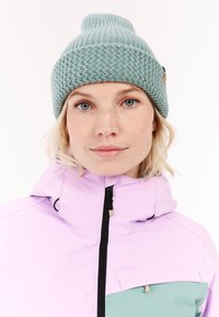 Giacca da sci viola chiaro con colletto alto, dotata di una tasca color teal. Berretto lavorato a maglia color teal con un motivo testurizzato e un piccolo bottone in marrone.