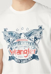 Bílé bavlněné tričko s modro-červeným potiskem grafiky s orlem, hvězdami a textem "Wrangler" a "Severní Karolína."