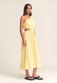 Keltainen, yhdellä olkapäällä oleva mekko, jossa on leikkausyksityiskohta, joustava vyötärö ja laskeutuva kangas. Malli käyttää mustia varvassandaaleja, joissa on litteä pohja.