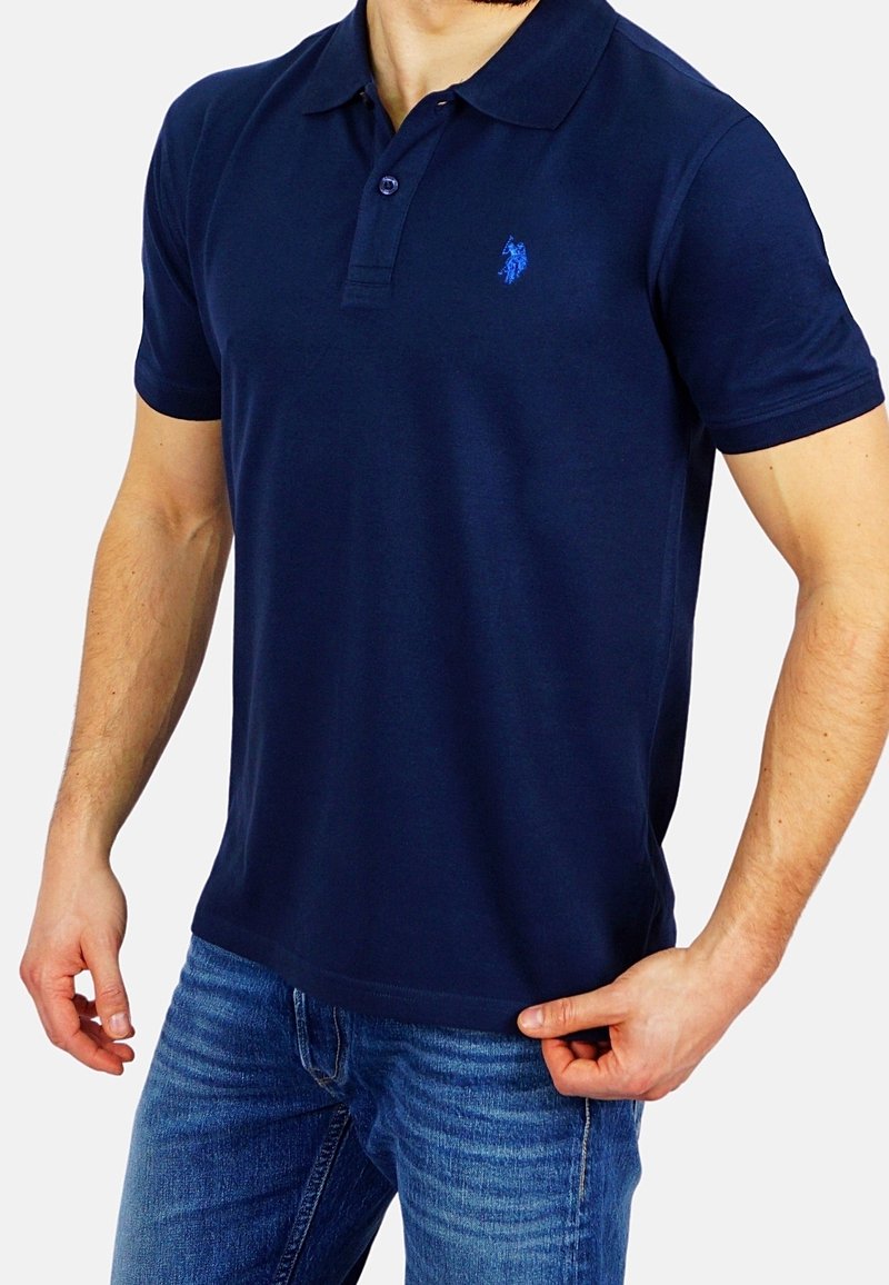 Homme portant un polo bleu marine à manches courtes et un jean bleu, tenant l'ourlet du polo d'une main.