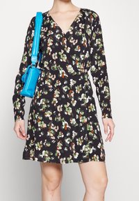 Czarna sukienka z długim rękawem w kwiatowy wzór w odcieniach zieleni, bieli i pomarańczy. Dekolt w kształcie litery V; zestawiona z małą niebieską torebką.