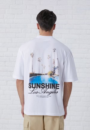 Bela bomba od bombaža s velikim grafičnim tiskom bazena in palm, na katerem je v debelem črnem napisu "SUNSHINE Los Angeles".