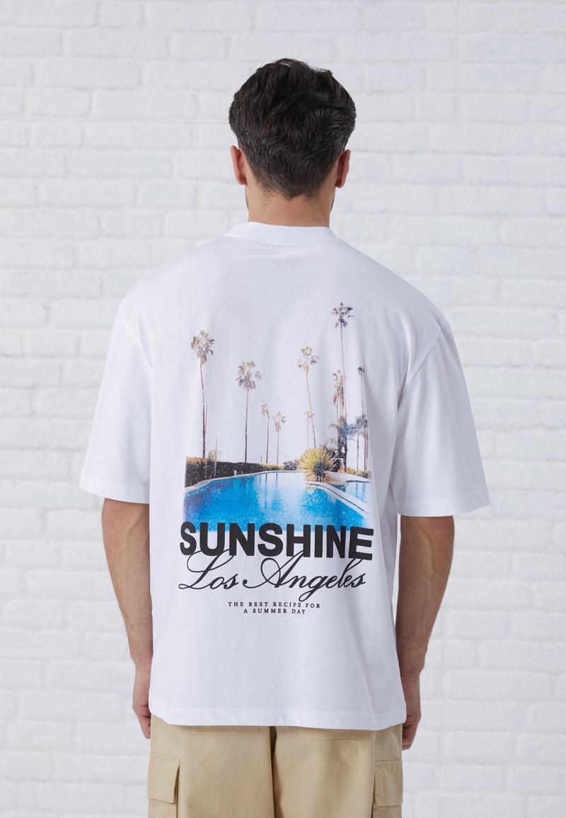 White cotton t-shirt featuring a large graphic print of a swimming pool and palm trees with the text "SUNSHINE Los Angeles" in bold black.