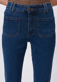 High-waisted blue jeans made from denim, featuring a smooth texture, copper button closure, and two back pockets with orange stitching.