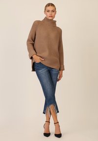 Maglione a collo alto a coste beige abbinato a jeans blu scuro con orli frangiati e tacchi neri, creando un look a strati.