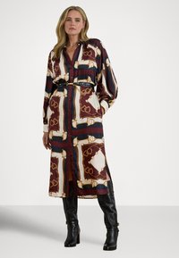 Γυναίκα που φοράει ένα μακρύ εμπριμέ φόρεμα με κουμπιά, ζώνη μαύρου χρώματος και μαύρα ψηλοτάκουνα μπότες μέχρι το γόνατο, στέκεται με το ένα χέρι στην τσέπη.