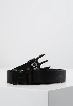 Černý tkaný pás s černou plastovou přezkou s označením "Calvin Klein Jeans", s bílým potiskem na popruhu, stočený na bílé ploše.