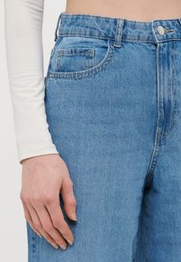 Ανοιχτό μπλε τζιν παντελόνι με ψηλή μέση, πέντε τσέπες και διακριτές ραφές. Λείο ύφασμα με άνετη εφαρμογή.