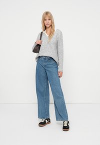 Jauna sieviete ar garām blondām matēm, ģērbusies pelēkā džemperī, plašās zilos džinsos, melnās un baltās sporta apavos, tur tumšu plecu somu.