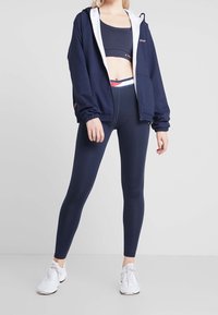 Kvinna i marinblå sport-bh, leggings, zip-up jacka och vita träningsskor som står mot en enkel bakgrund.