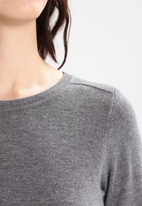 Pull gris en maille avec un col rond, une texture douce et un détail aux épaules. Le tissu semble léger et légèrement extensible.