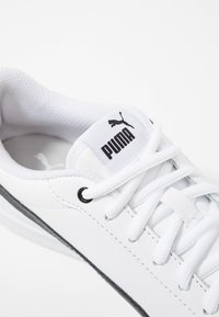 Gros plan d'une chaussure de sport Puma blanche avec un logo noir sur la languette, des lacets blancs et un détail de bande noire sur le côté.
