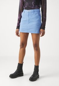 Jupe en denim bleu clair mini avec poches avant, fermeture à bouton et détails de couture visibles, associée à des bottines noires.