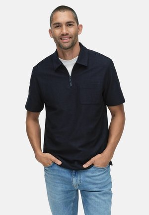 Korte ærmer navy polo shirt med lynlåskrave, med en brystlomme og en glat tekstur. Båret over en hvid t-shirt og parret med blå jeans.