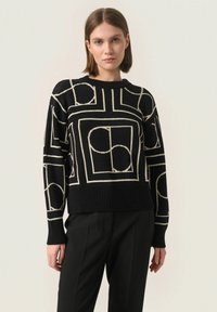 Czarny sweter dzianinowy z geometrycznym wzorem w kolorze kremowym. Okrągły dekolt, długie rękawy i ściągacz u dołu. Noszony z czarnymi spodniami.