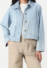 Ljusblå jeansjacka med vertikala vita ränder, spetsig krage, två framfickor och knappstängning. Korthanterad längd med fransade kanter.