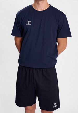 Mężczyzna ubrany w granatowy t-shirt hummel z krótkim rękawem i pasujące krótkie spodenki, stoi z rękami splecionymi za plecami na białym tle.