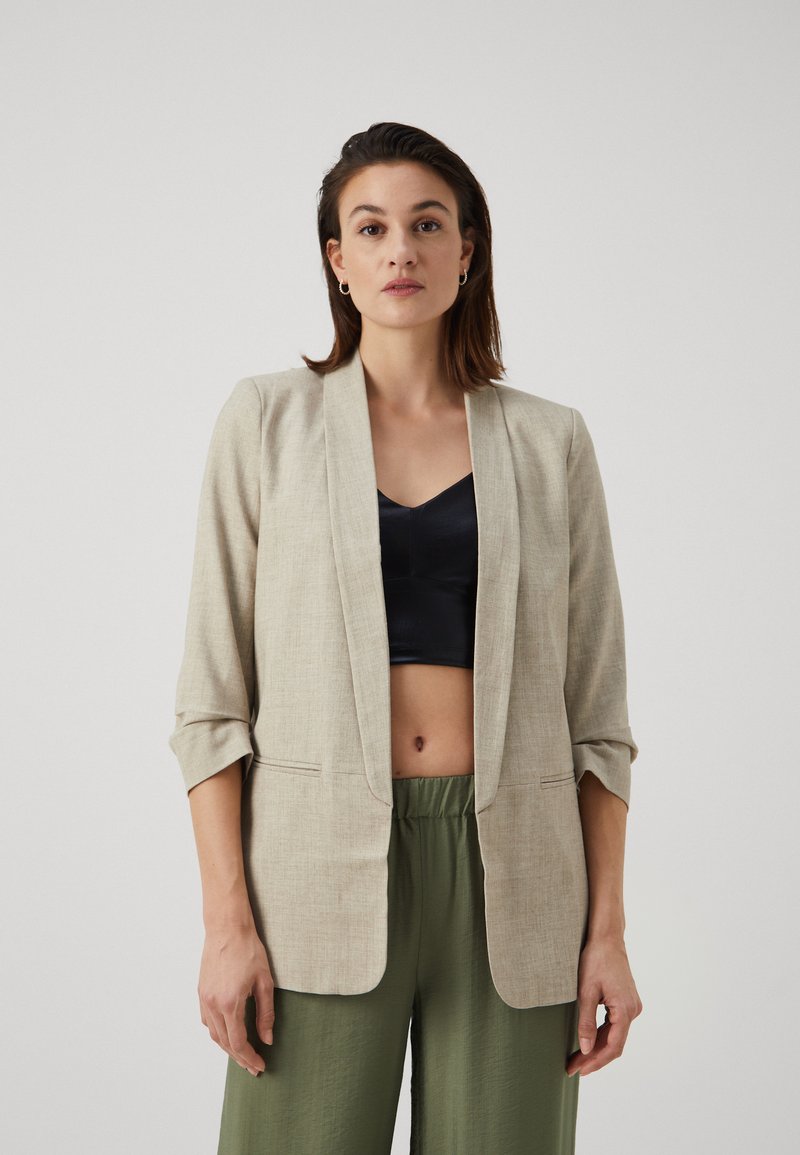 Blazer en lin beige avec des revers et des manches trois-quarts, associé à un haut noir et un pantalon vert. Texture lisse, design simple.