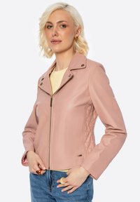 Giacca in ecopelle rosa con zip frontale, pannelli laterali trapuntati e colletto a pressione. Indossata sopra un top giallo, abbinata a jeans blu.