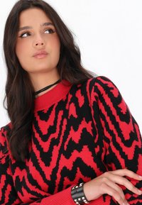 Pulover tricotat roșu și negru, cu un model în zigzag, guler rotund și manșete cu nervuri; purtat cu o brățară neagră cu tivuri.