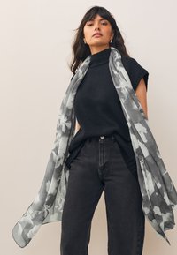 Rento musta neulepusero yhdistettynä korkeavyötäröisiin mustiin farkkuihin, jossa harmaa kukkakuvioinen huivi hulmuaa hartioilla.